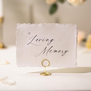 May include: A white card with the words "In Loving Memory" in elegant black script. The card is held by a gold wire holder, suitable for a memorial or wedding event. The card has a deckled edge.