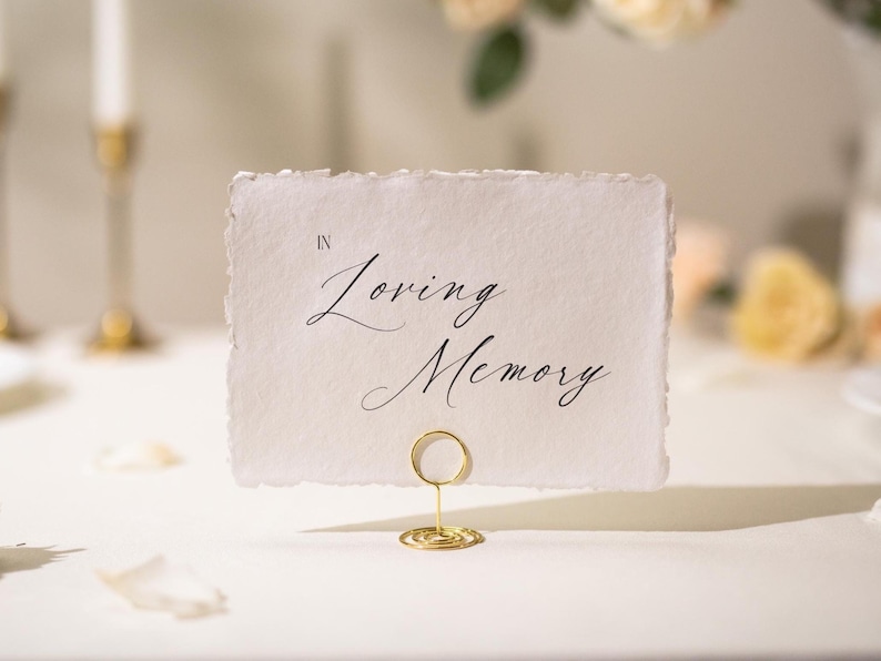 May include: A white card with the words "In Loving Memory" in elegant black script. The card is held by a gold wire holder, suitable for a memorial or wedding event. The card has a deckled edge.