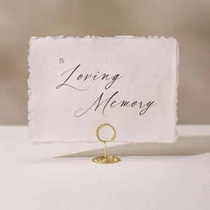 May include: A white card with the words "In Loving Memory" in elegant script is held by a gold wire holder. The card has a deckled edge, and the background is a soft, neutral color.