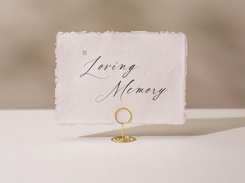 May include: A white card with the words "In Loving Memory" in elegant script is held by a gold wire holder. The card has a deckled edge, and the background is a soft, neutral color.