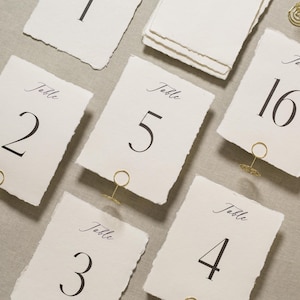 May include: Off-white table number cards with torn edges, featuring elegant black numbers and the word "Table" in script. Each card is held by a gold wire holder. The numbers 1, 2, 3, 4, 5, and 16 are visible.