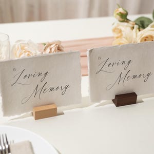 May include: Two rectangular cards with the text "Loving Memory" in elegant script, displayed on wooden blocks. The cards are on a white surface, with floral arrangements and glassware in the background. The cards are for a memorial or wedding.