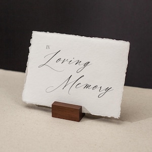 May include: A white card with the words "In Loving Memory" in elegant script is displayed on a small, dark wooden stand. The card has a textured, deckled edge. The background is a neutral beige and black.