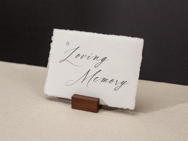 May include: A white card with the words "In Loving Memory" in elegant script is displayed on a small, dark wooden stand. The card has a textured, deckled edge. The background is a neutral beige and black.
