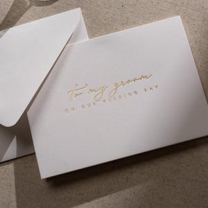 May include: A white card with gold foil lettering that reads "To my groom on our wedding day".