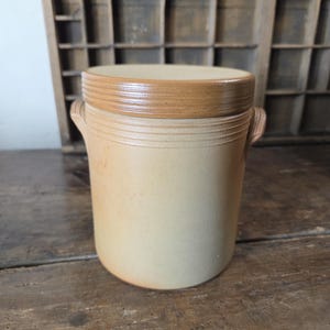 Puede incluir: Una vasija de gres cilíndrica con cuerpo de color beige claro y una tapa lisa de color marrón que contrasta. El recipiente cuenta con dos pequeñas asas laterales y detalles de ranuras horizontales alrededor del borde superior y la tapa.