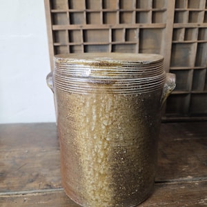 Puede incluir: Una vasija de gres cilíndrica con un acabado de esmalte salino en tonos marrón y bronceado moteado, que presenta una tapa plana, dos pequeñas asas laterales y detalles acanalados horizontales alrededor del borde superior, con textura.