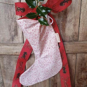 Peut inclure: Une chaussette de Noël blanche ornée d'un motif rouge de formes festives. Elle est suspendue à un ruban rouge décoré de cerfs et d'écureuils. Du houx et des baies ornent le haut de la chaussette.