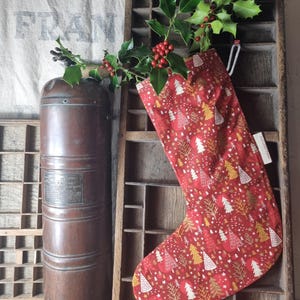 Peut inclure: Une chaussette de Noël rouge avec un motif d'arbres festifs, remplie de houx et de baies, est suspendue dans une étagère en bois. Un récipient en cuivre vintage se trouve à gauche. L'arrière-plan comprend un sac en toile de jute avec du texte délavé.