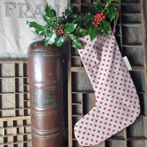 Peut inclure: Une chaussette de Noël avec un motif d'étoiles rouges, remplie de houx et de baies rouges. Elle est suspendue à côté d'un récipient en métal vintage et d'un casier d'imprimeur en bois. Le fond présente un texte estompé.