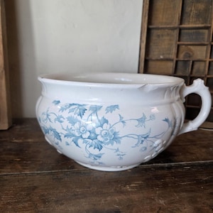 Può includere: Un vaso da notte in ceramica bianca con un motivo floreale blu di viticci e foglie. Il recipiente ha un corpo arrotondato, un manico laterale singolo e una piccola sbeccatura sul bordo superiore. Il design include fiori e foglie dettagliati.