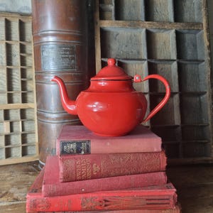 Könnte beinhalten: Eine leuchtend rote Teekanne mit Deckel steht auf einem Stapel alter roter Bücher. Im Hintergrund befinden sich ein hoher, kupferfarbener Kanister und ein Holzorganisator. Die Szene hat eine Vintage-Ästhetik.