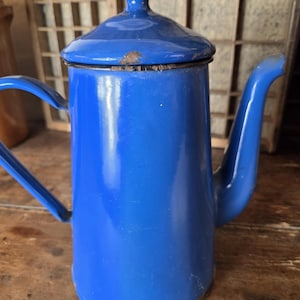 Könnte beinhalten: Eine blaue Emaille-Kaffeekanne im Vintage-Stil mit Deckel und Griff. Die Kanne hat einen Ausguss und einen Griff an der Seite. Der Deckel hat einen kleinen Knopf oben. Die Kanne weist Gebrauchsspuren auf, mit etwas Rost.