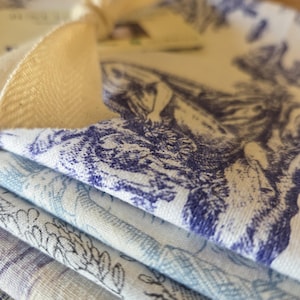 Peut inclure: Une pile de serviettes en tissu pliées présentant un motif toile de Jouy bleu et blanc avec des illustrations détaillées de personnages et de feuillage. Les serviettes sont attachées par un ruban crème et incluent une petite étiquette en papier.