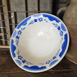 May include: A white ceramic bowl with a blue floral design around the rim. The bowl has a scalloped edge and is set on a wooden surface. The blue pattern includes stylized flowers and leaf motifs.