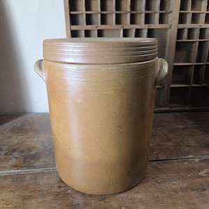 Peut inclure: Un pot en grès cylindrique de couleur beige avec un couvercle assorti doté de rainures horizontales et deux petites anses latérales. Le récipient présente une finition lisse et uniforme ainsi qu'une structure verticale robuste et classique.