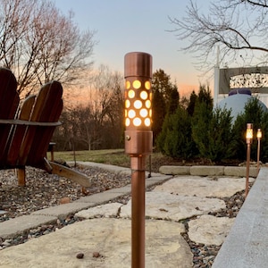 May include: A copper-colored outdoor path light featuring a cylindrical head with a perforated pattern that emits a warm, glowing light. The fixture is mounted on a slender, matching copper-colored metal stake, standing upright in a garden setting.