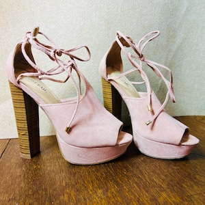 May include: A pair of pale pink platform heels with open toes and wooden block heels. The heels have a lace-up design and gold-coloured accents. The shoes are displayed on a wooden surface.