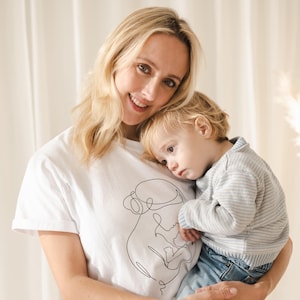 May include: A woman wearing a white t-shirt with a black line drawing of a mother and child. She is holding a young child wearing a gray striped sweater and blue jeans. The background is a soft, neutral color.