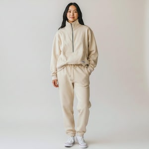 May include: A beige tracksuit consisting of a quarter-zip sweatshirt and matching sweatpants. The sweatshirt has a high collar and a black zipper. The sweatpants have a drawstring waist. The outfit is worn with white sneakers.