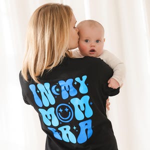 May include: Black t-shirt with the text "IN MY MOM ERA" in blue bubble letters, a smiley face, and stars. A baby is being held by a person wearing the shirt.