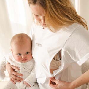 May include: A white t-shirt with a zipper for easy breastfeeding access. The shirt has the word "mamie" embroidered on the front. A baby is being held by the person wearing the shirt.