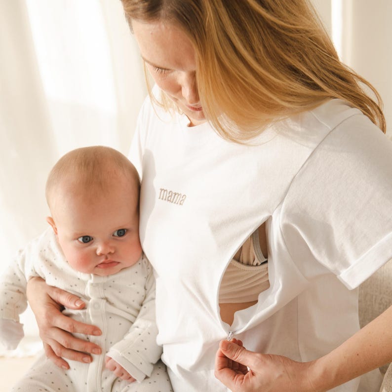 May include: A white t-shirt with a zipper for easy breastfeeding access. The shirt has the word "mamie" embroidered on the front. A baby is being held by the person wearing the shirt.