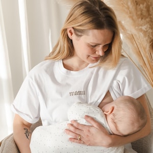 May include: A woman wearing a white t-shirt with the word "mama" embroidered on the front is breastfeeding a baby. The baby is wearing a white and gray polka dot onesie.