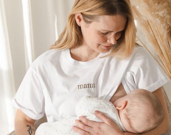 May include: A woman wearing a white t-shirt with the word "mama" embroidered on the front is breastfeeding a baby. The baby is wearing a white and grey polka dot onesie.