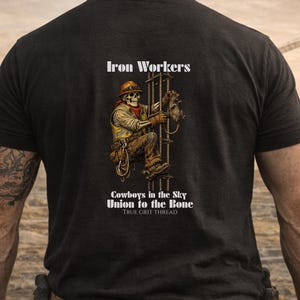 Pu&ograve; includere: T-shirt nero con un'immagine di un operaio edile scheletrico che scala una struttura in acciaio. Il design include il testo "Iron Workers", "Cowboys in the Sky" e "Union to the Bone". La maglietta ha anche il testo "TRUE GRIT THREAD."