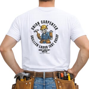 Può includere: T-shirt bianca con grafica di uno scheletro carpentiere e la scritta "Union Carpenter Skilled Labor Isn't Cheap". Lo scheletro indossa un elmetto e abiti da lavoro, con un martello e chiodi. Visibile una cintura porta attrezzi in pelle marrone.
