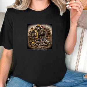 Può includere: T-shirt nera con grafica di scheletri di operai edili, la scritta "Spoiled Blue Collar Wife" e "True Grit Thread". La grafica include un cantiere, attrezzi e un casco giallo.