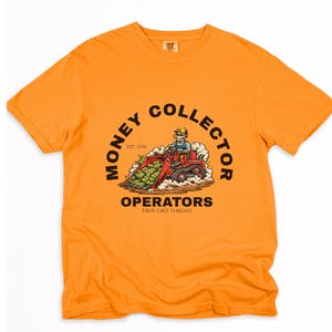 Puede incluir: Camiseta naranja con un gráfico que muestra un esqueleto operando una cargadora frontal roja llena de dinero. El texto dice "MONEY COLLECTOR" y "OPERATORS" con "EST. 1896" y "TRUE GRIT THREAD" debajo.