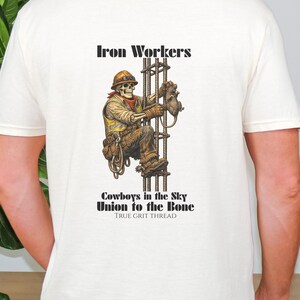Pu&ograve; includere: T-shirt color crema con una grafica dettagliata di un operaio edile scheletrico che scala una struttura in acciaio. Il design include il testo "Iron Workers", "Cowboys in the Sky" e "Union to the Bone".
