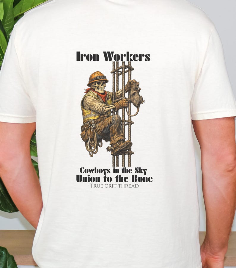 Pu&ograve; includere: T-shirt color crema con una grafica dettagliata di un operaio edile scheletrico che scala una struttura in acciaio. Il design include il testo "Iron Workers", "Cowboys in the Sky" e "Union to the Bone".