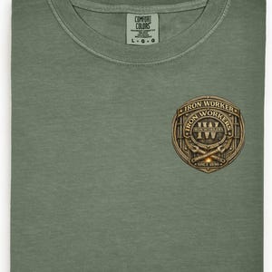 Pu&ograve; includere: T-shirt verde oliva con scollo tondo. Presenta un emblema dorato e marrone con le scritte "Iron Workers" e "Since 1896". L'etichetta riporta "Comfort Colors".