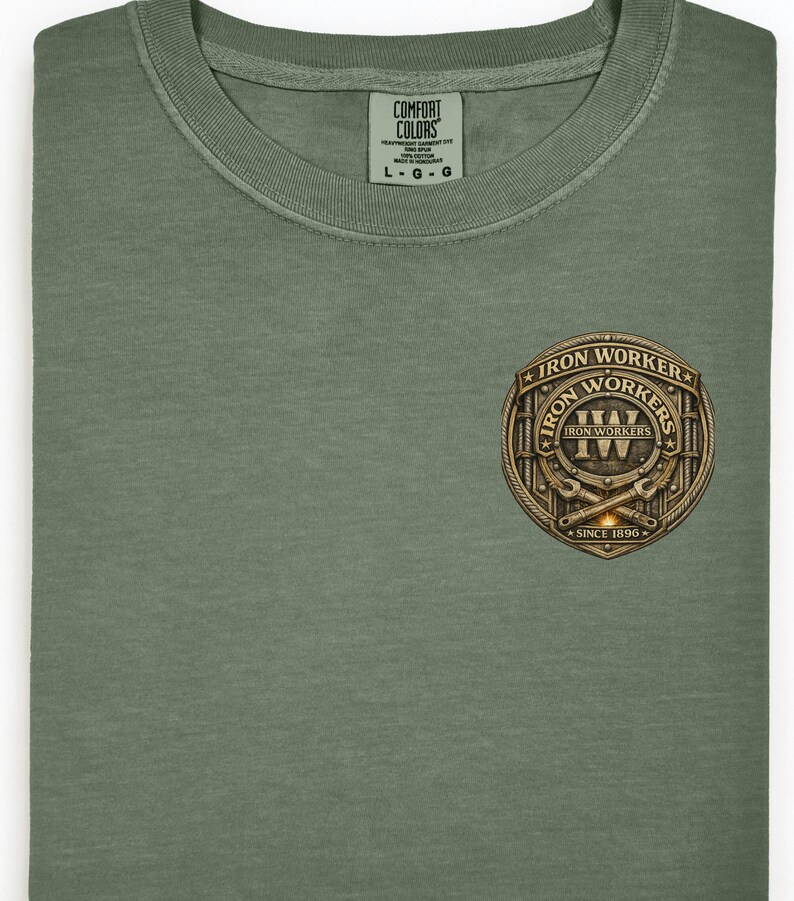 Pu&ograve; includere: T-shirt verde oliva con scollo tondo. Presenta un emblema dorato e marrone con le scritte "Iron Workers" e "Since 1896". L'etichetta riporta "Comfort Colors".