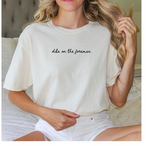 Può includere: T-shirt color crema con la scritta "dibs on the foreman" in corsivo nero. La maglietta ha una vestibilità ampia e sembra essere realizzata in materiale morbido. La modella indossa pantaloncini bianchi.