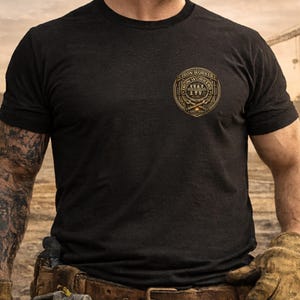 Pu&ograve; includere: T-shirt nero girocollo con un emblema dorato e marrone degli Iron Workers sul petto. L'emblema presenta il testo "Iron Workers" e attrezzi incrociati. La maglietta &egrave; realizzata in un tessuto scuro e strutturato.