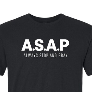 May include: A black short-sleeved t-shirt featuring white text on the chest that reads A.S.A.P in a bold, sans-serif font, with the words ALWAYS STOP AND PRAY printed in a smaller, thinner white font directly underneath the main acronym.