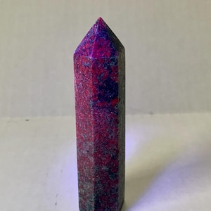 May include: A tall, hexagonal crystal point with a vibrant colour palette. The crystal features a deep red base with speckled dark blue and purple hues, culminating in a pointed tip. The polished surface reflects light, highlighting its natural beauty.