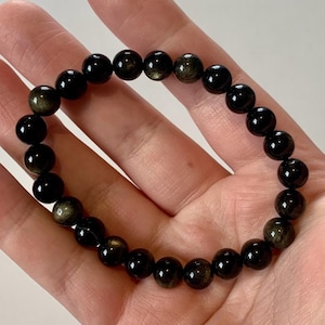 May include: A black beaded bracelet with a golden sheen. The bracelet is made of round, polished beads strung together. The beads have a glossy finish and reflect light, creating a shimmering effect. The bracelet is shown on a light background.