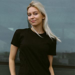 Puede incluir: Una persona con una camiseta negra de manga corta. La persona tiene el pelo largo y rubio y está sonriendo. El fondo es una superficie texturizada de color azul claro.