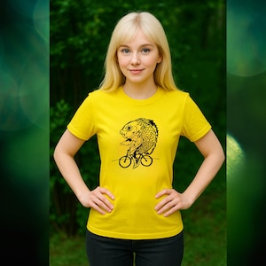 Peut inclure: Un t-shirt jaune avec une illustration noire d'un poisson à vélo. Le poisson est détaillé et placé au centre. Le t-shirt est à manches courtes et en tissu doux. Le fond est vert flou.