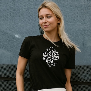 Może przedstawiać: Czarny t-shirt z krótkim rękawem z białym nadrukiem graficznym na piersi. Wzór składa się z trzech ułożonych w stos falistych wstęg z cyrylicą, otoczonych małymi ikonami w kształcie pigułek oraz rozproszonymi kropkami, tworzącymi wyraźny motyw.