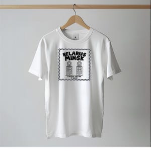 May include: White t-shirt with a black and white graphic featuring the text "Belarus Minsk" and illustrations of two buildings. The graphic is framed by a checkered border, with "Limited Edition T-Shirt" text below.