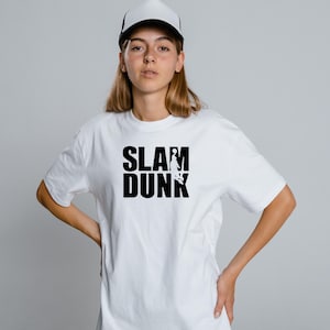 Può includere: T-shirt bianca con la scritta "SLAM DUNK" in nero. Il design include la silhouette di un giocatore di basket. La persona nell'immagine indossa un berretto da baseball bianco e nero.