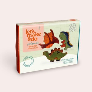 Puede incluir: Una caja rectangular para un kit de manualidades de dinosaurios de madera para pintar. El empaque muestra ilustraciones de un pterodáctilo rojo, un T-Rex verde y un estegosaurio verde. El texto incluye let's make & do y QR code included.