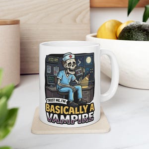Puede incluir: Taza de cerámica blanca con un esqueleto de dibujos animados con uniforme azul, sentado en un escritorio con un monitor de ordenador y una lámpara. La taza tiene el texto "Trust me, I'm basically a vampire."