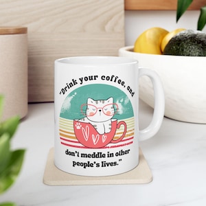 Puede incluir: Taza de café de cerámica blanca con una ilustración de gato con gafas dentro de una taza roja. La taza tiene el texto "Drink your coffee, and don't meddle in other people's lives."